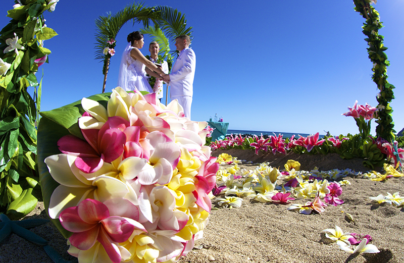 Pink and yellow plumeria bouquet in front of a couple exchanging vows under a green arch on the beach